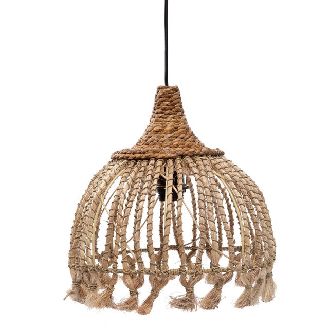 A bohemian-style woven rattan pendant light with a tassel fringe, hanging in a warm, sunlit Mediterranean-style living room with a white sofa, textured pillows, and potted olive trees.