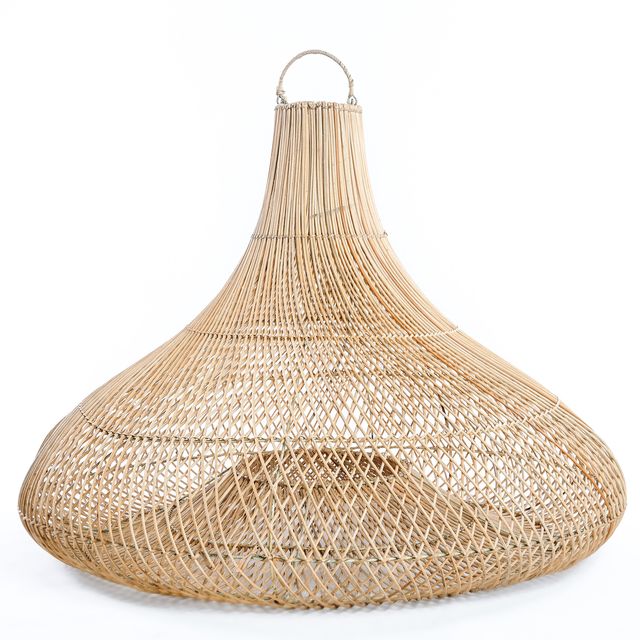 A woven natural fiber pendant light hangs over a round wooden dining table with wicker chairs on a covered patio. The scene includes potted olive trees, a vase with greenery, and a stone wall background with trees visible outside.