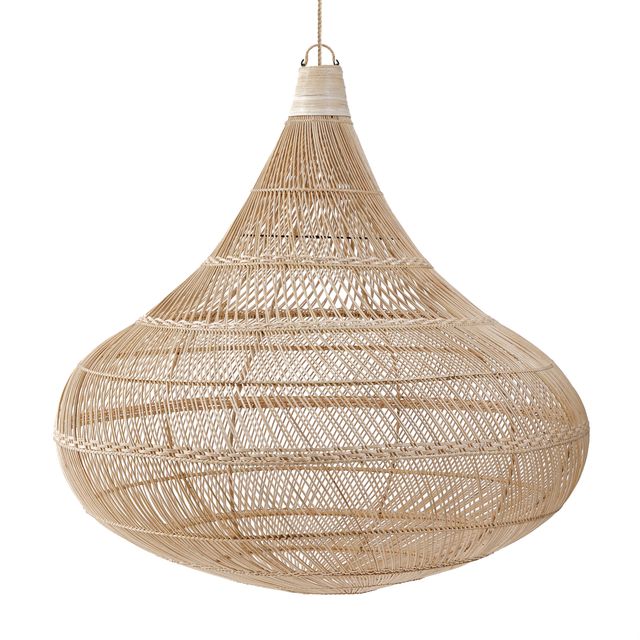 A large woven wicker pendant light hangs over a rustic wooden dining table on an outdoor patio with a sea view background.