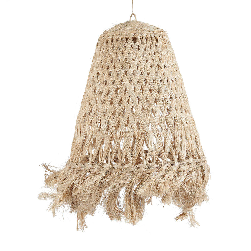 A woven natural-fiber pendant lamp with fringe hangs over a rustic stone table in an outdoor patio setting with olive trees, plants, and wooden beams.