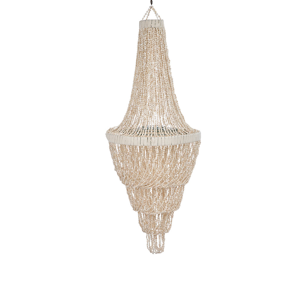 A large chandelier made from shells hangs over a rustic wooden dining table with wicker chairs in an outdoor patio setting with greenery and a covered wooden ceiling.